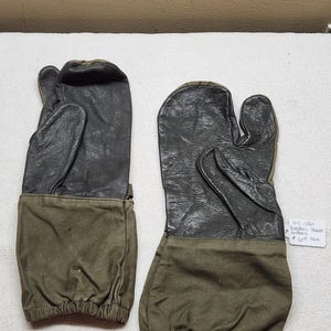 Puede incluir: Un par de manoplas de color verde oliva y negro con una palma y un pulgar de cuero oscuro texturizado unidos a un puño de lona. Una pequeña etiqueta blanca está unida a la manopla derecha con el texto escrito a mano U.S. COLD WEATHER TRIGGER MITTENS.