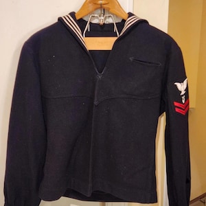 May include: A dark navy blue long-sleeve sailor jumper featuring a traditional wide collar with white trim, a V-neckline, and a white eagle emblem above two red chevron stripes on the left sleeve. The cuffs also feature white trim detailing.