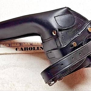 May include: A black leather holster with a wrap-around strap and brass-colored metal rivets. The holster features the embossed text "Schützenregiment 1914". A white ruler is positioned underneath, showing the holster is approximately 8 inches long.