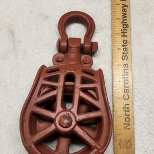Puede incluir: Un bloque de polea de metal de color marrón rojizo con un marco de celosía y un grillete giratorio en la parte superior. Una regla de madera marcada North Carolina State Highway Patrol está junto a la polea, mostrando una longitud de 19 cm.