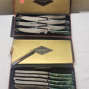May include: A set of silver-toned knives with marbled green handles, presented in two rectangular boxes labeled Community Plate. The knives feature metallic blades and decorative handles, arranged neatly within the lined storage cases.