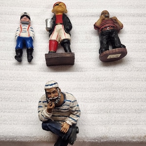 May include: Four hand-painted resin figurines of sailors and pirates. One figure wears a striped shirt and blue pants, another is a pirate with an eye patch, a third holds a sack, and the fourth is a seated sailor in a striped shirt and blue pants.