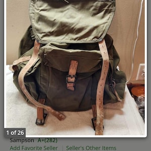 May include: An olive green canvas rucksack featuring a large top flap, a front pocket with a metal buckle closure, and light tan leather straps. The rucksack includes a drawstring closure under the main flap and integrated metal frame components.