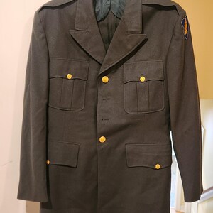 May include: An olive drab military-style service jacket featuring a notched lapel, four front pockets with buttoned flaps, and gold-toned buttons. The jacket includes shoulder epaulets and a blue and yellow patch on the left sleeve.