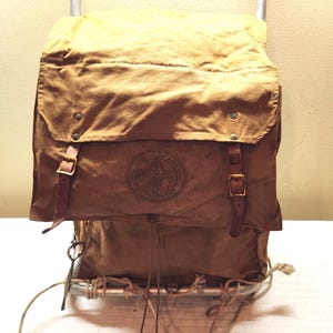 May include: A tan canvas backpack with a metal frame, featuring two brown leather straps with brass buckles and a circular Boy Scouts of America logo stamped on the front flap. The pack includes a drawstring closure and hanging rope ties.