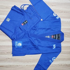 Könnte beinhalten: Ein königsblauer brasilianischer Jiu-Jitsu-Gi-Anzug, bestehend aus einer langärmeligen Jacke und einer passenden Hose. Die Jacke ist mit dem RVCA-Logo auf der Brust und den Ärmeln versehen, während die Hose einen Kordelzug in der Taille aufweist.