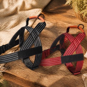 May include: Two dog harnesses, one black and one red, featuring reflective silver stripes, adjustable straps, a brown leather-like top handle, and a black metal D-ring for leash attachment. The harnesses have a structured design with padded sections.