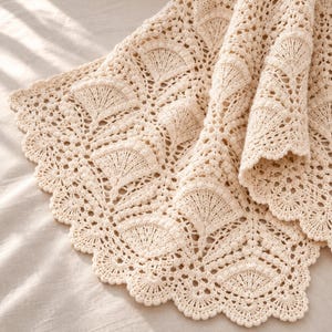 May include: A cream-colored crochet blanket featuring an intricate fan-shaped stitch pattern throughout the fabric. The edges of the textile are finished with a scalloped border, showcasing a detailed, repetitive design with small, raised bobble accents.