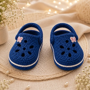 May include: A pair of dark blue crochet baby shoes featuring a white trim along the sole, a rounded toe with six circular cutouts, and a small pink and white butterfly appliqué on the outer side of each shoe, complete with a functional heel strap.
