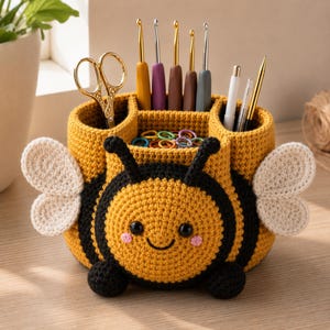May include: A crocheted bee-shaped desk organizer in yellow and black with white wings, featuring multiple compartments holding gold scissors, various colored crochet hooks, pens, and a collection of small plastic stitch markers inside.