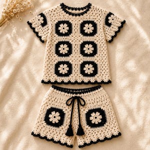 Puede incluir: Un conjunto de ganchillo de dos piezas que consta de una parte superior de manga corta y pantalones cortos. Ambas piezas están hechas de hilo crema y negro, con un motivo cuadrado repetitivo con un diseño de flor central y borde festoneado.