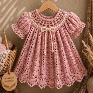 May include: A dusty pink crochet dress featuring a round neckline with a grid-like pattern, short puff sleeves with scalloped edges, and a flared skirt with vertical textured stripes. A cream-colored cord is tied into a bow at the center chest.
