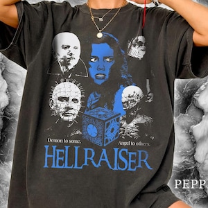 May include: A charcoal gray short-sleeve t-shirt featuring a blue and white graphic print of characters from the movie Hellraiser, including a puzzle box and the text "Demon to some. Angel to others. HELLRAISER." The shirt has a crew neckline.