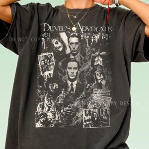 May include: A charcoal gray short-sleeve t-shirt featuring a black and white collage graphic from the film Devil's Advocate. The design includes multiple character portraits, a city skyline, and the text Devil's Advocate printed at the top.