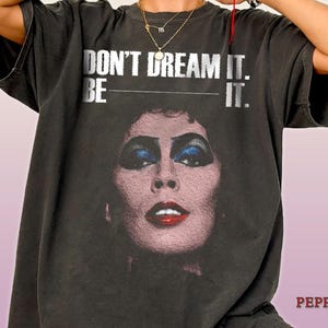 Op de afbeelding: Een antracietgrijs T-shirt met korte mouwen en een grafische print van een gezicht met opvallende make-up, inclusief blauwe oogschaduw en rode lippenstift. Boven het gezicht staat de witte tekst: DON'T DREAM IT. BE IT. Het shirt heeft een ronde hals.