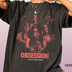 Op de afbeelding: Een antracietgrijs t-shirt met korte mouwen en een rode grafische print van vier gezichten en de tekst "OBSESSION A film by CURRY BARKER". Het ontwerp bevat kleine rode hart- en sterpictogrammen verspreid boven de gezichten op de voorzijde.