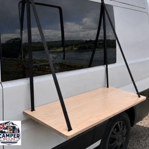 May include: A rectangular light-coloured wooden shelf mounted to the exterior side of a white van. The shelf is supported by two black straps that attach to the top of the window frame. A logo with the text CAMPER BUILDS .CO.UK is in the corner.