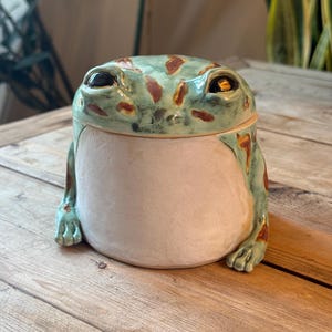 Pode incluir: Um pote de cerâmica em forma de sapo com uma tampa verde-clara com manchas castanhas e dois olhos escuros. A base do pote é um cilindro bege liso e mate com duas pernas de sapo esculpidas que se estendem pelos lados.