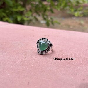 May include: A silver ring featuring a smooth, triangular-shaped green stone set in a decorative metal frame with small bead accents. The text Shivjewels925 is visible on the surface near the ring. The metal has a polished, oxidised finish.