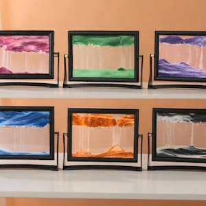 May include: Six rectangular sand art frames with black borders, each mounted on a rotating stand. The frames contain layers of coloured sand in pink, green, purple, blue, orange, and black, creating shifting landscape patterns within the glass.