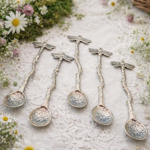 May include: Five silver-toned metal spoons with handles shaped like textured tree branches, each topped with a dragonfly figure. The bowl of each spoon features a hammered, dimpled surface texture, creating a reflective and uneven appearance.