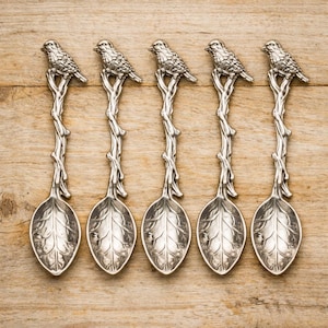 May include: A set of five silver-toned metal spoons featuring a handle shaped like a textured tree branch with a small bird perched at the top and a bowl designed to resemble a detailed leaf with visible veins and a pointed tip.