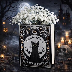 May include: A rectangular black planter shaped like a book titled Book of Spells Magick Cat Energy, featuring a central illustration of a black cat with green eyes and moon phases. The planter is filled with a bouquet of small white flowers.