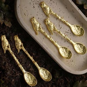 Puede incluir: Un juego de cinco cucharas de metal dorado, cada una con un mango texturizado en forma de rama con un pequeño pájaro posado cerca del extremo. Los cuencos de las cucharas tienen una superficie martillada irregular sobre una bandeja beige.