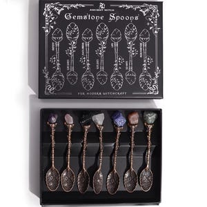 Könnte beinhalten: Ein Set aus sieben kupferfarbenen Metalllöffeln mit floralen Griffen und rohen Edelsteinaufsätzen, präsentiert in einer schwarzen Schachtel mit der Aufschrift Ancient Witch Gemstone Spoons. Der Deckel zeigt weiße Strichzeichnungen der Löffel.