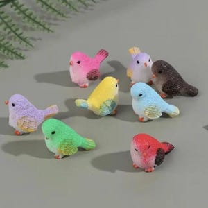 May include: A collection of eight small bird figurines in various colours including pink, yellow, light blue, green, red, purple, and brown. Each miniature bird features a textured surface, a small orange beak, and a rounded body shape.