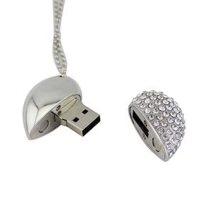 May include: A silver-toned heart-shaped USB flash drive pendant on a ball chain. The drive is separated into two pieces, with the exposed metal connector on one side and a removable cap encrusted with clear, sparkling rhinestones on the other.