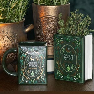 Könnte beinhalten: Ein dunkelgrüner Keramikbecher und ein passender rechteckiger Übertopf in Buchform, beide mit dem Text "GREEN WITCH Book of Spells" sowie botanischen und himmlischen Illustrationen. Dahinter stehen zwei bronzefarbene Töpfe mit Kräutern.