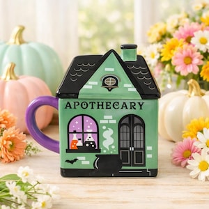 Puede incluir: Una taza de cerámica verde claro con forma de casa, techo negro y asa morada. El frente muestra la palabra APOTHECARY, una ventana con frascos de pociones, un caldero negro, un pequeño murciélago y un diseño de puerta doble en negro.
