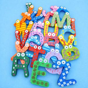 May include: A collection of colorful wooden alphabet letters, each featuring a pair of cartoon eyes and unique patterns such as stripes, polka dots, swirls, and floral motifs. The letters are arranged in a pile against a solid light blue background.