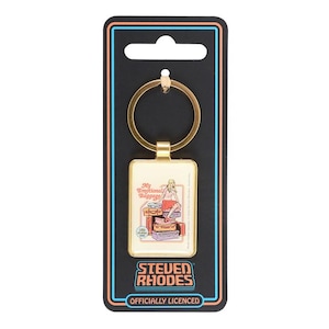 May include: A rectangular gold-toned metal keychain featuring a graphic illustration of a person sitting on a stack of suitcases with the text My Emotional Baggage. The keychain is attached to a black display card with the text STEVEN RHODES.