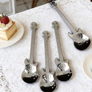 Pode incluir: Quatro colheres de metal prateado em forma de guitarras elétricas. Cada colher apresenta um braço longo como cabo e um corpo de guitarra que serve como concha, com detalhes gravados da escala e da ponte na superfície polida.