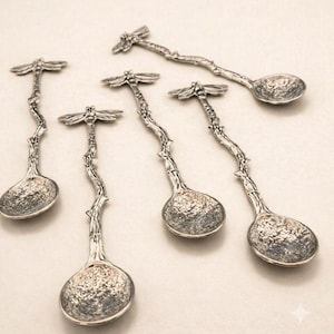 May include: A set of five silver-toned metal spoons featuring a textured, branch-like handle and a dragonfly motif at the top. Each spoon has a round, hammered-finish bowl, creating a cohesive set of decorative cutlery with a rustic, organic design.