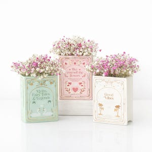 Puede incluir: Tres jarrones rectangulares de cerámica con forma de libro llenos de flores de paniculata rosas y blancas. El jarrón izquierdo es verde claro con el texto Myths, Fairy Tales & Legends. El central es rosa con el texto Buy yourself the flowers. El derecho es blanco con el texto Good Vibes.