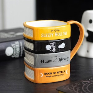 May include: A ceramic mug shaped like a stack of four books with an orange handle. The spines are labeled The Legend of Sleepy Hollow, Ghost Stories with a ghost icon, The Haunted Library, and Book of Spells with crescent moon icons.