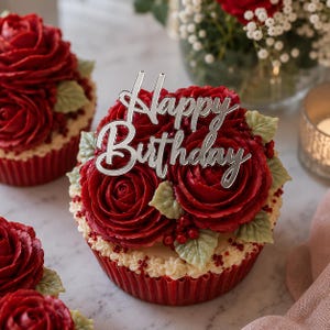 May include: A cupcake topped with deep red buttercream roses, green leaves, and small red berries, featuring a silver-colored acrylic cake topper with the text Happy Birthday in a cursive script, set in a dark red pleated paper baking cup.