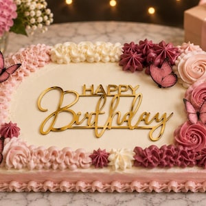 Puede incluir: Un pastel rectangular con glaseado blanco, decorado con remolinos de crema de mantequilla rosa y rosa oscuro, además de varios recortes de mariposas rosas. Un adorno acrílico dorado en el centro muestra el texto Happy Birthday.