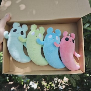 Puede incluir: Cuatro peluches con forma de criaturas estilizadas con orejas redondeadas y cuerpos curvos. Los peluches son de color verde menta, verde lima, azul brillante y rosa. Cada uno presenta dos ojos circulares simples y una boca pequeña.