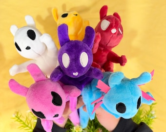May include: A collection of six small plush toys in various colors including white, yellow, purple, red, pink, and blue. Each creature features large, distinct eyes, pointed ears, and a soft, fuzzy texture, arranged in a cluster.