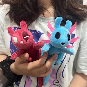 May include: Two plush toys featuring stylized creature designs. One toy is deep red with a white eye patch and a white eye, while the other is bright blue with large black eyes and pink felt gills extending from the sides of its head.