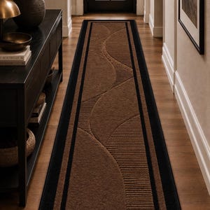 Puede incluir: Una alfombra de pasillo rectangular larga con un centro marrón oscuro y un borde negro. La alfombra muestra un patrón repetitivo de líneas curvas y secciones texturizadas con crestas paralelas, creando un diseño geométrico.