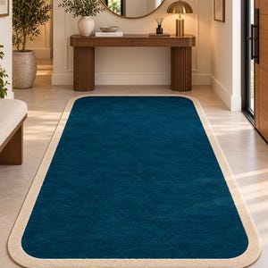 May include: A rectangular area rug featuring a deep teal blue center with rounded corners and a wide, solid beige border. The rug has a low-pile texture and is positioned flat on a light-colored floor, extending from the foreground into the room.