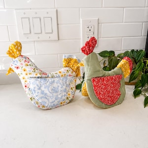 May include: Two fabric chicken-shaped kitchen accessories. The left one features a white base with a yellow and pink floral pattern and a light blue floral pocket. The right one is sage green with a red floral wing and a red comb.