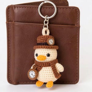 May include: A crochet duck keychain featuring a cream-colored body, orange feet and beak, a brown scarf, and a brown top hat adorned with a small clock face. The duck holds a round pocket watch accessory with Roman numerals on its right side.