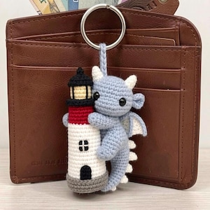 May include: A crochet keychain featuring a light blue dragon hugging a lighthouse. The lighthouse has a white base, a red middle section, and a black top with a yellow light. The dragon has small white horns, black eyes, and a braided loop attachment.