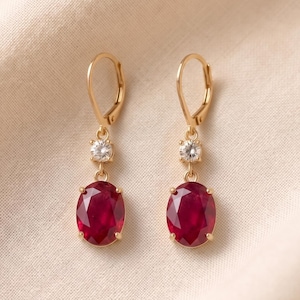 Könnte beinhalten: Ein Paar goldene Ohrhänger mit Klappbügel, bestehend aus einem facettierten, oval geschliffenen, tiefroten Rubin, der unter einem einzelnen, rund geschliffenen, klaren weißen Diamanten hängt. Die Fassung besteht aus poliertem Gold.
